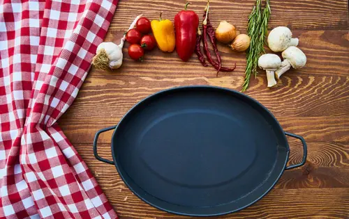 A pizza pan on bare wood beside a red gingham cloth, peppers, garlic and mushrooms
